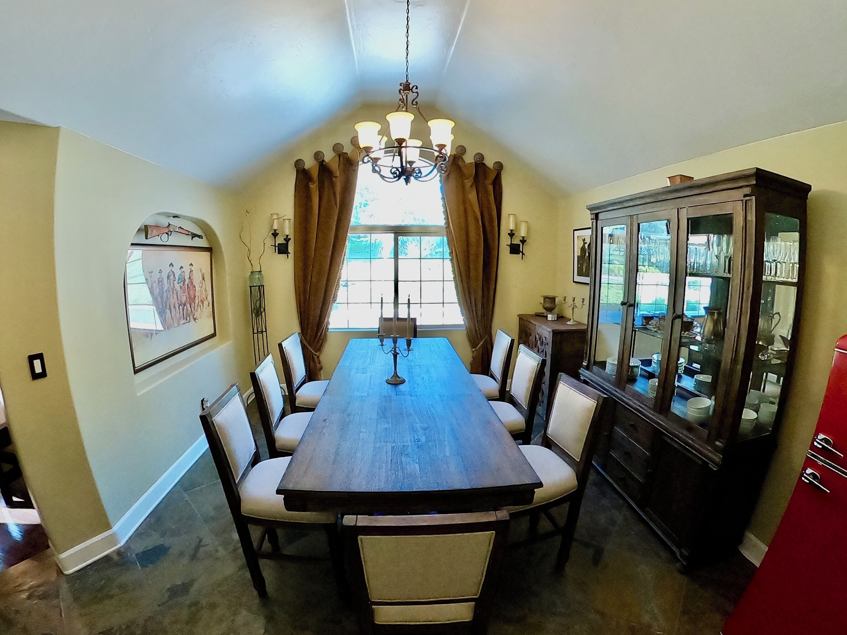 Une salle à manger avec une longue table en bois, six chaises, un lustre, un buffet aux portes vitrées et un tableau accroché au mur.