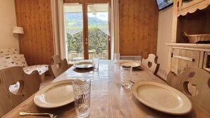 A dining area with a wooden table set for two, a television on the wall, and a view of greenery outside.