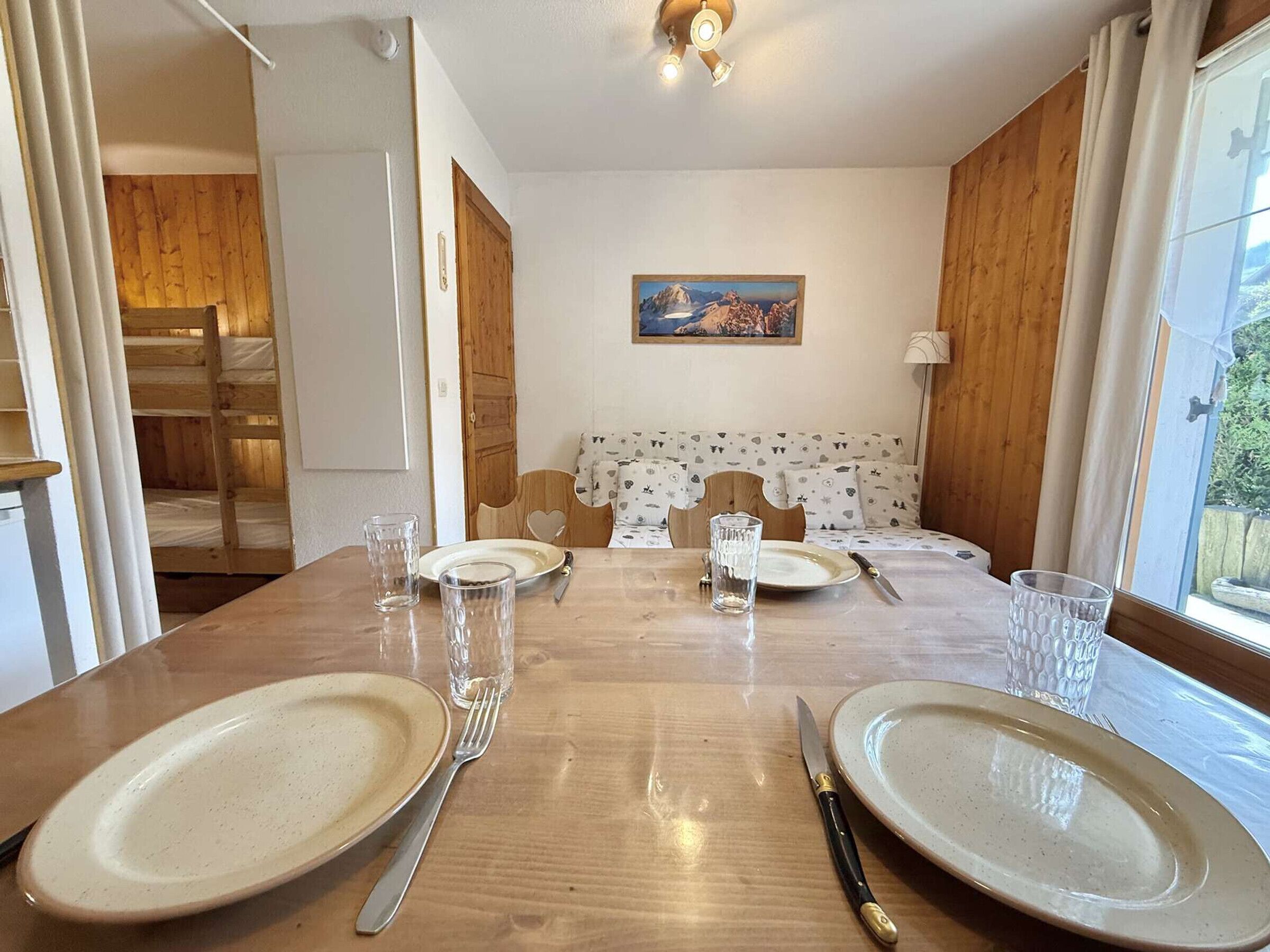 Un comedor con una mesa de madera preparada para dos personas, dos platos, dos tenedores, dos cuchillos y dos vasos. Hay un cuadro en la pared y una ventana con cortinas.