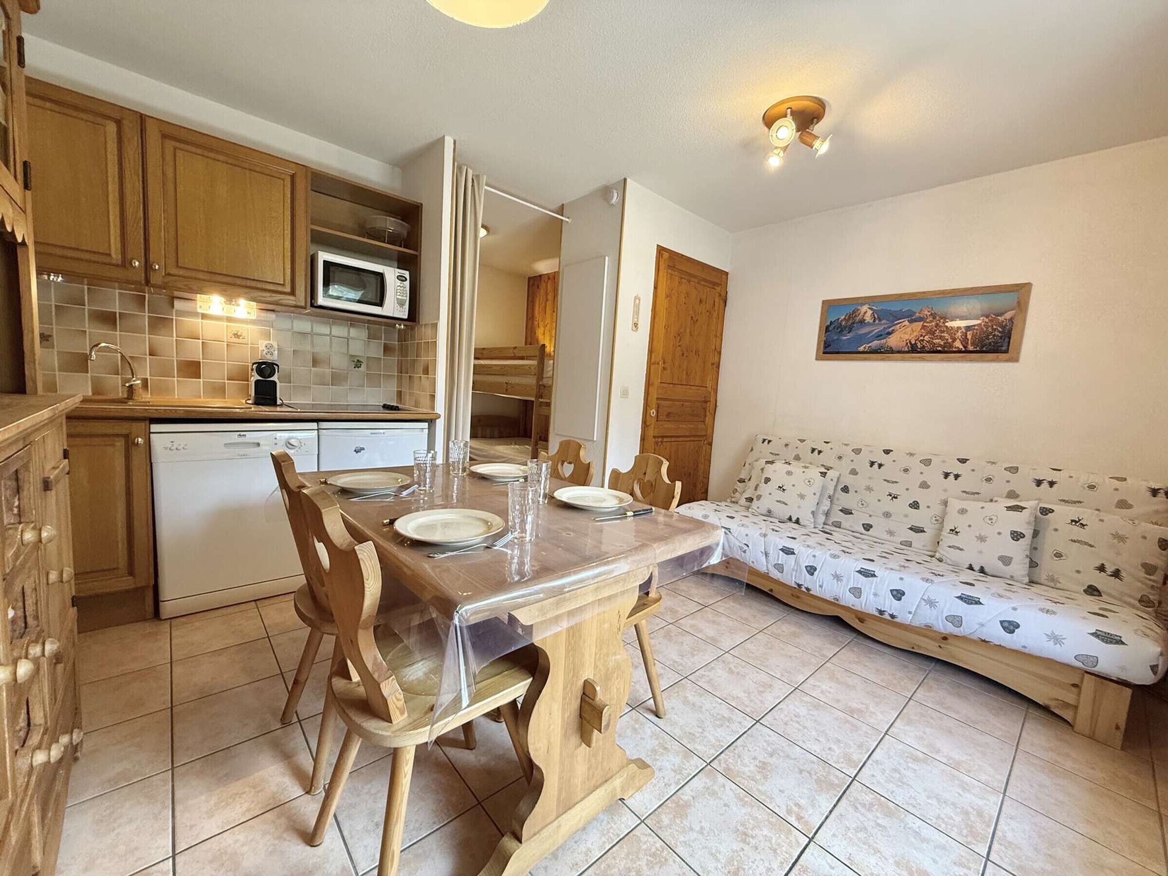 A compact kitchen with wooden cabinets, a microwave, and a dishwasher. A dining table with chairs is set for four. A sofa with a patterned cover is against the wall.