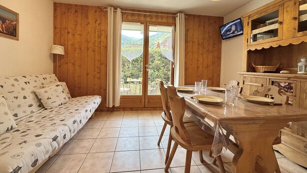 A cozy room with a wooden sofa, a dining table with chairs, a TV, and a view of the outdoors.