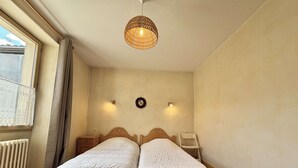 A bedroom with two single beds, a wooden headboard, a small bedside table, a wall-mounted light, a radiator, and a window with curtains. - Cozy apartment in Praz-sur-Arly, 2 rooms, near village (Praz-sur-Arly)