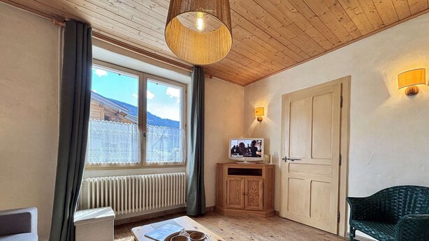 A cozy room with a wooden ceiling, a window with a view of a building, a wooden cabinet with a television, a bench with a tray, and a green chair. - Cozy apartment in Praz-sur-Arly, 2 rooms, near village (Praz-sur-Arly)