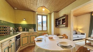 A cozy dining area with a round table set for four, wooden cabinets, a window with a view, and a bookshelf. - Cozy apartment in Praz-sur-Arly, 2 rooms, near village (Praz-sur-Arly)