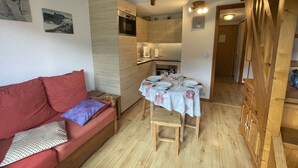 Uma sala de estar acolhedora com um sofá vermelho, mesa de jantar de madeira para quatro pessoas e uma cozinha com armários de madeira e forno embutido.