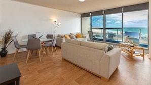 A modern living room with a large window, a sofa, a dining table with chairs, and a TV on a stand. - Oceanic 3 - On The Beach! (Forster)