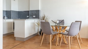A modern kitchen with white cabinets, a black tiled backsplash, and a dining area with a glass table and grey upholstered chairs. - Oceanic 3 - On The Beach! (Forster)