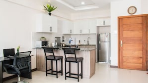 Una cocina moderna con armarios blancos, un refrigerador de acero inoxidable, un microondas y una cafetera. Hay dos taburetes de bar y una computadora portátil sobre un escritorio.
