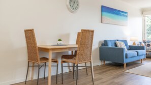 Um quarto com uma mesa de jantar de madeira, duas cadeiras de vime, um sofá azul e um quadro com uma cena de praia.