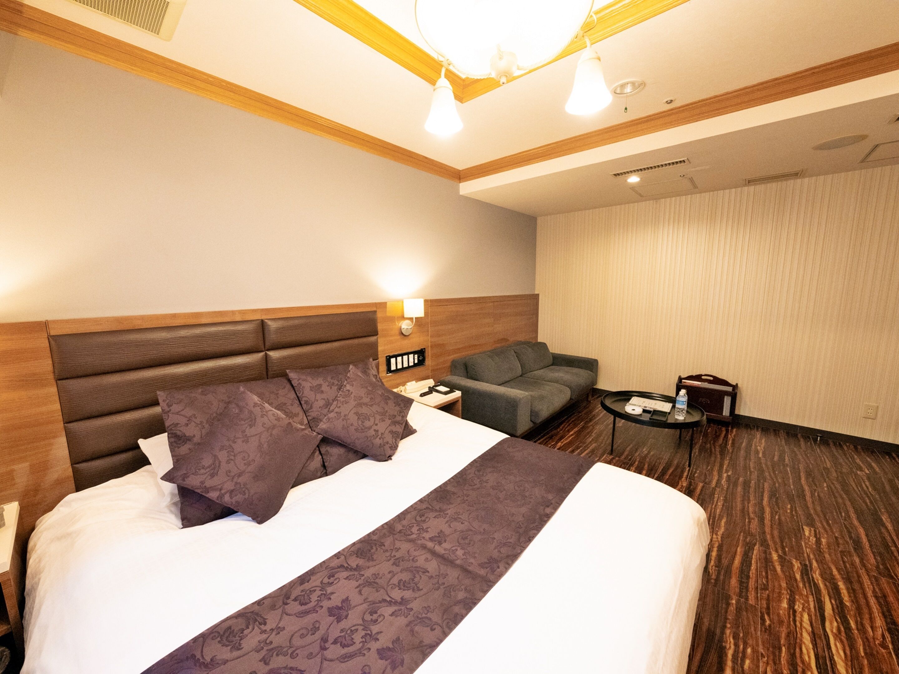 A hotel room with a large bed, a sofa, a small table, and a wall-mounted lamp.
