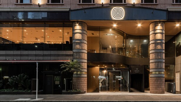 Fachada de hotel con diseño moderno, destacando un logotipo sobre las puertas, amplios ventanales de vidrio y una estructura de columna central.