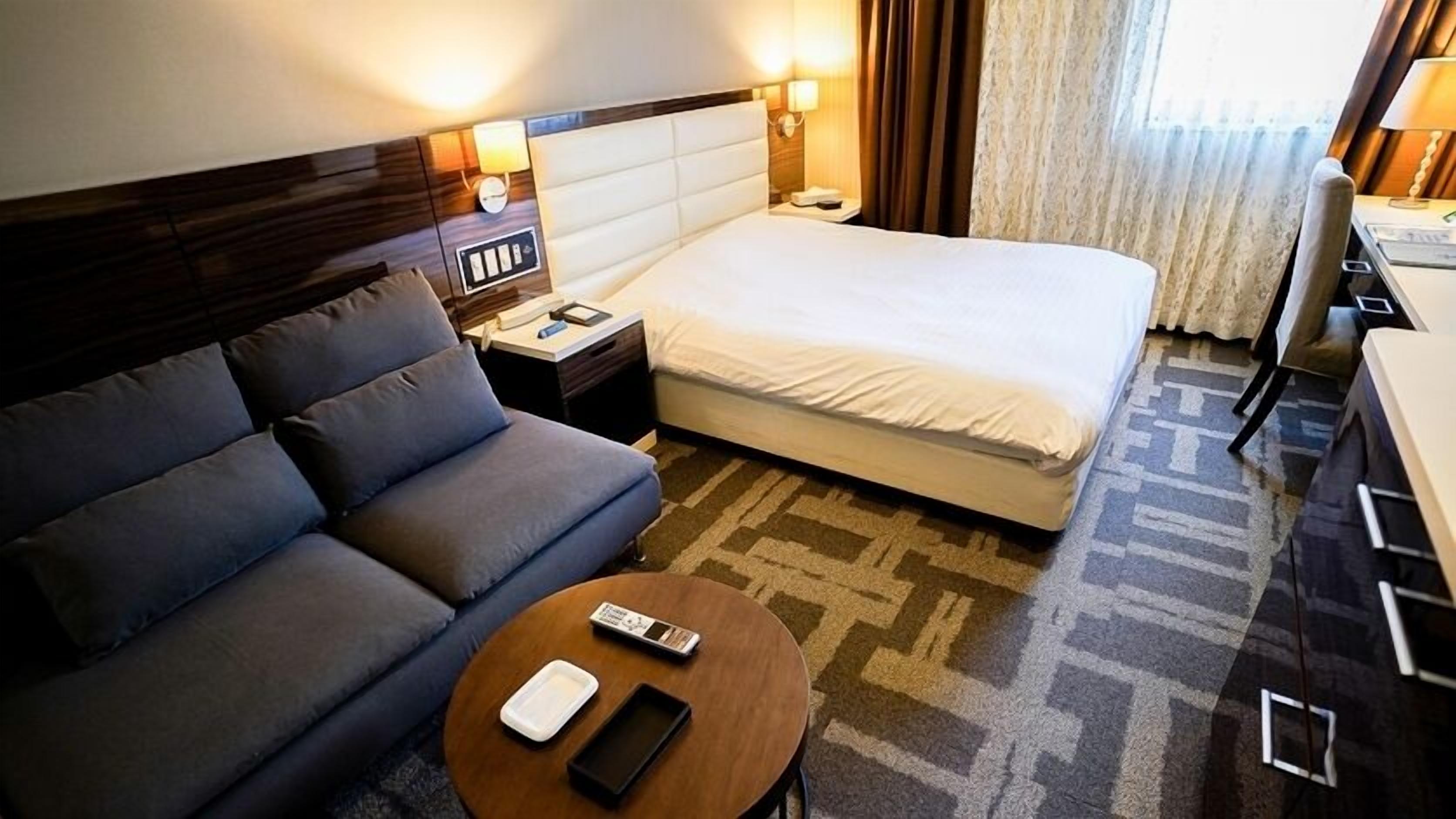 A hotel room with a bed, a sofa, a bedside table, a lamp, and a small table with a remote control.