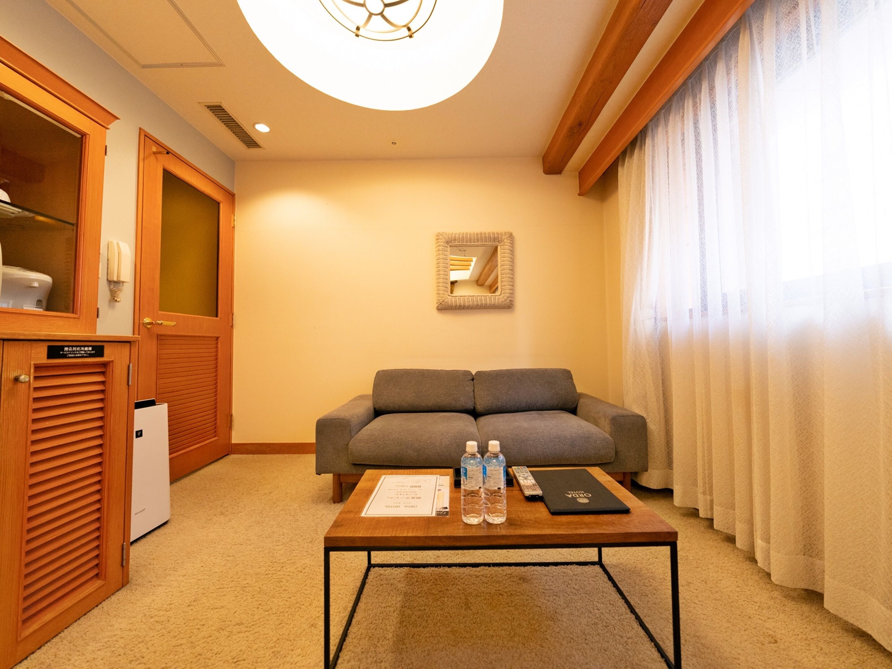 A hotel room with a grey sofa, a wooden cabinet, a small table with a remote control, a framed picture, and a window with sheer curtains.