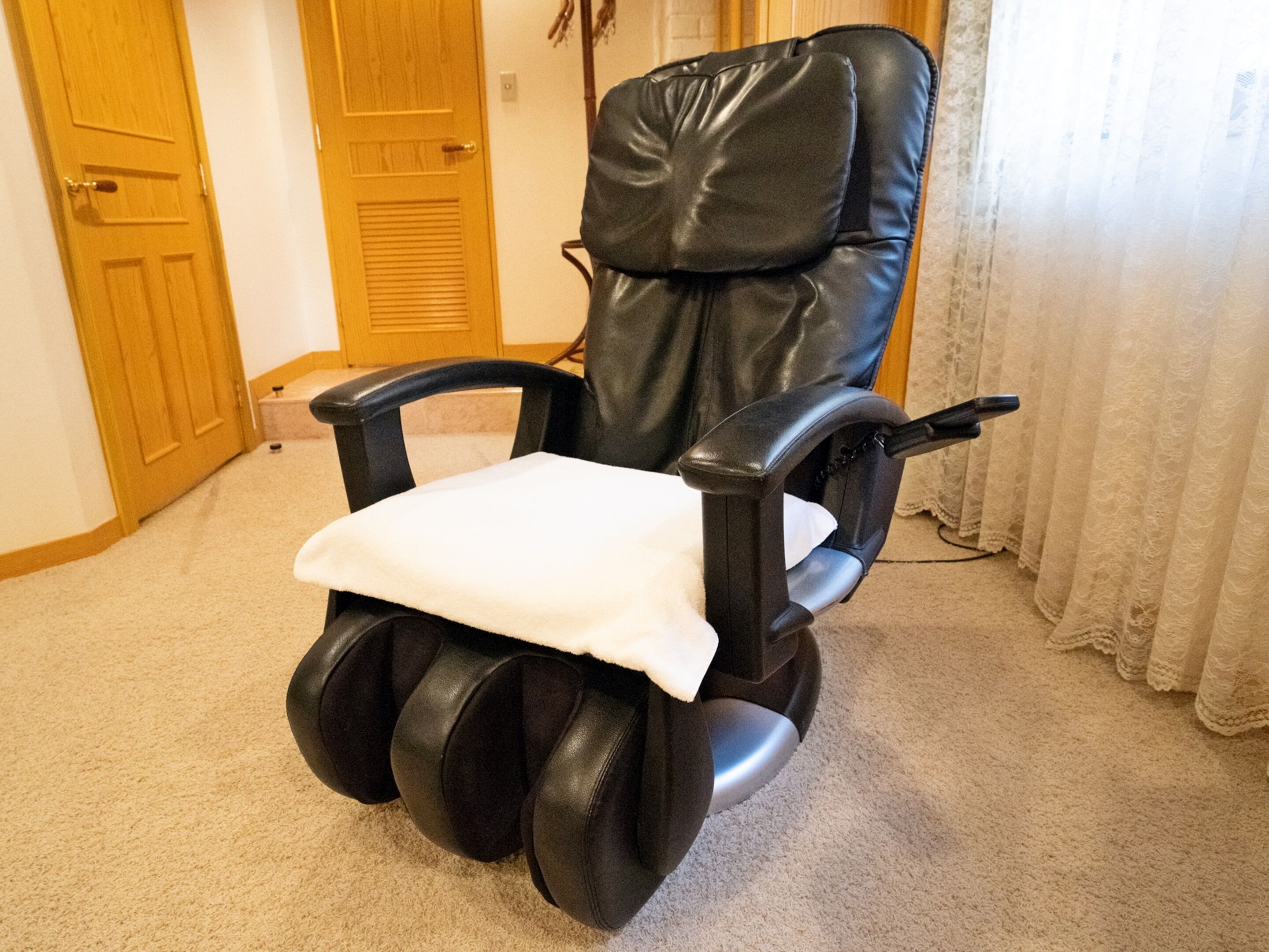 En massagefåtölj med fotstöd och en vit kudde i ett rum med beige matta och trädörrar.