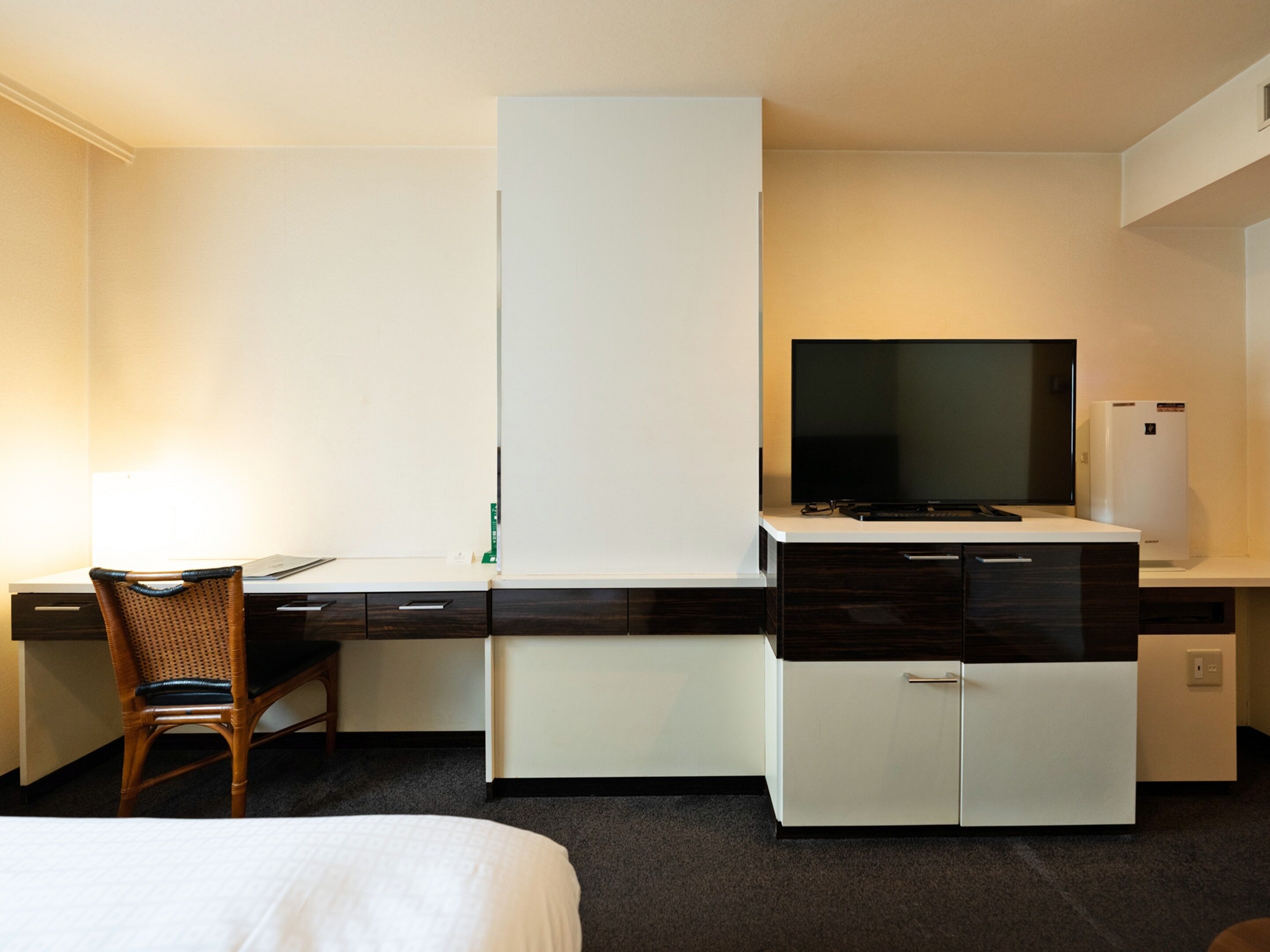 A hotel room with a bed, a desk with a chair, a flat-screen TV, a mini-fridge, and a wardrobe.