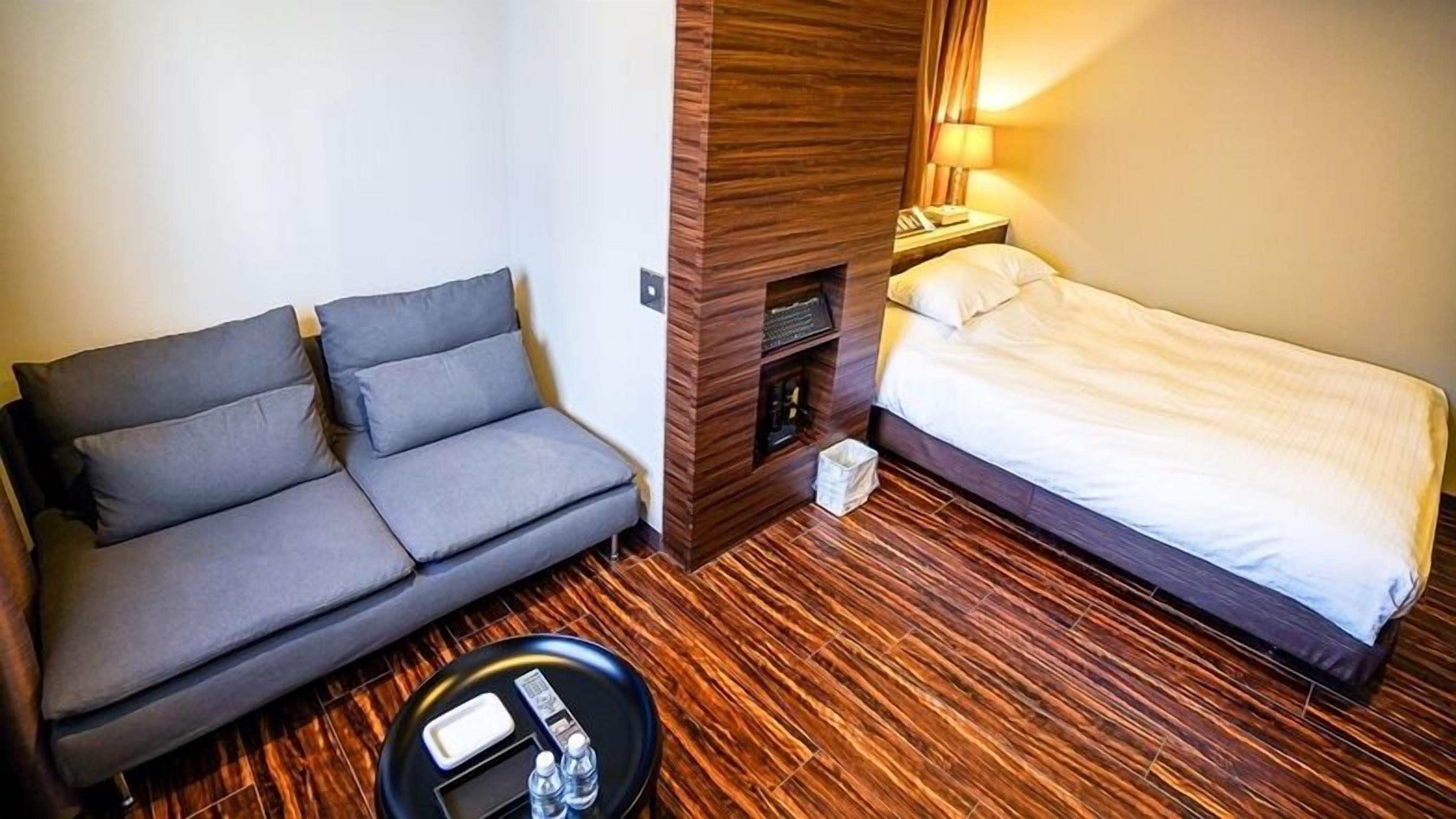A compact hotel room with a sofa, a single bed, a small table with a bottle and a tissue box, and a wooden accent wall.