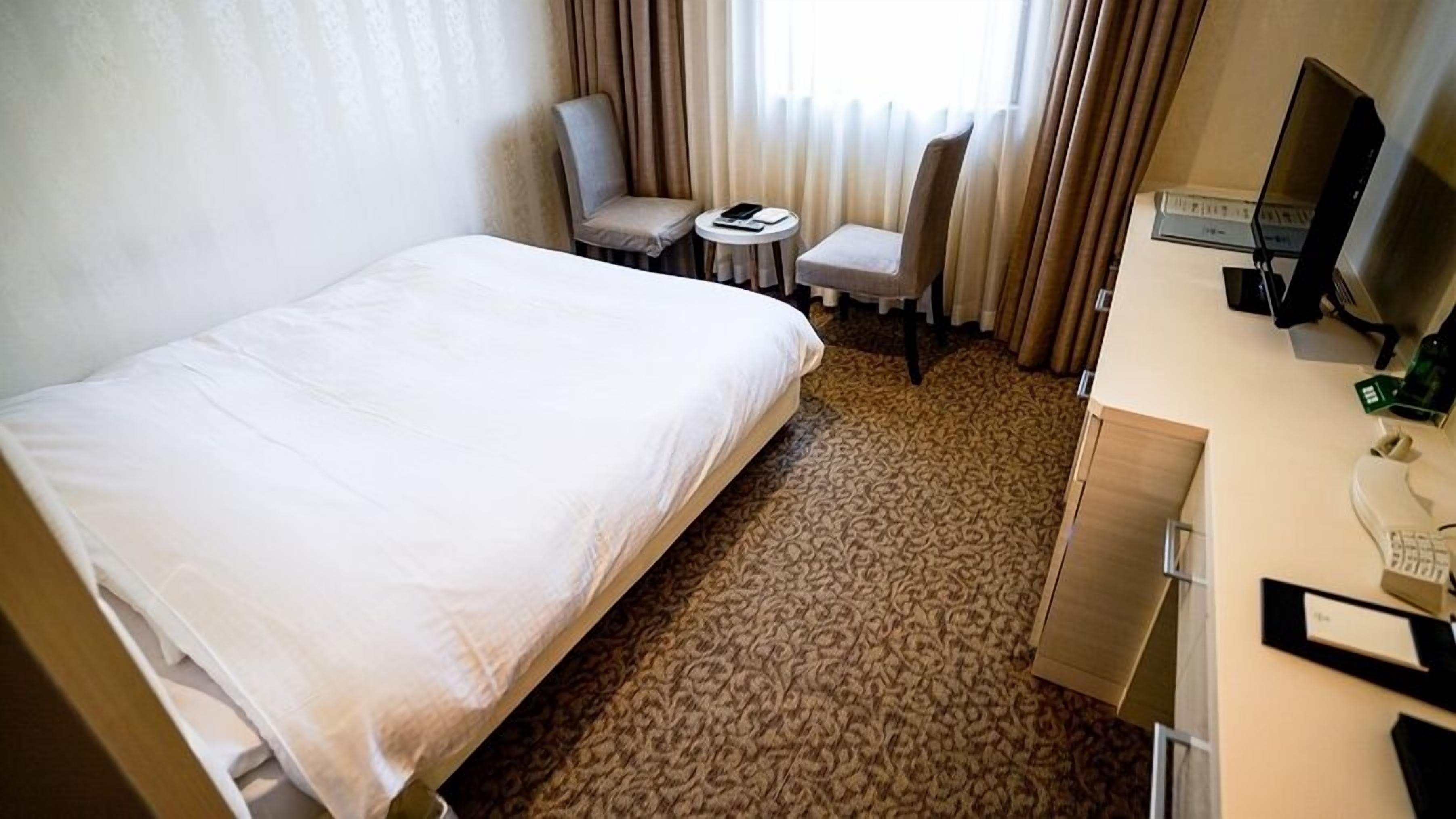 A hotel room with a bed, a desk with a computer, a chair, a small table with a book, and a window with curtains.