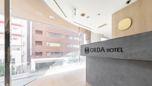 Un área de recepción con diseño moderno, que incluye un mostrador de recepción con el logotipo de ORDA HOTEL, grandes ventanales con vistas a la calle y un techo de madera.