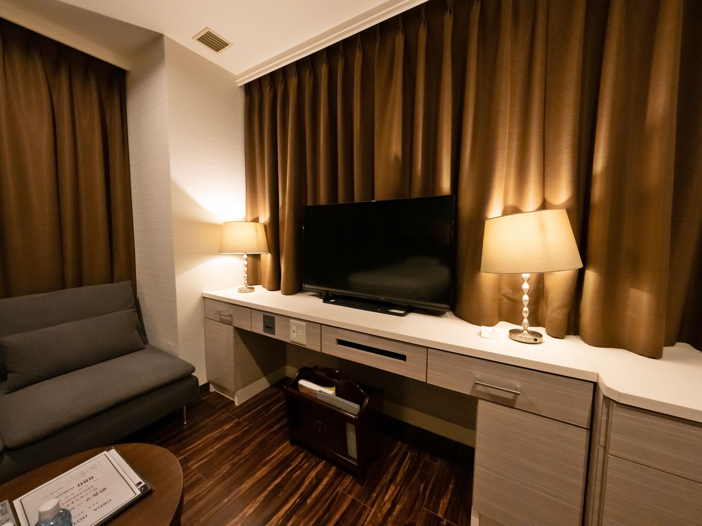 A hotel room with a flat-screen TV mounted on the wall, a sofa, a desk with a lamp, and a wooden floor.