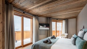 A bedroom with a wooden ceiling, a large bed with a grey blanket, a TV on the wall, and a balcony with wooden railings. - Exclusive Chalet in Courchevel Village (Courchevel 1550)