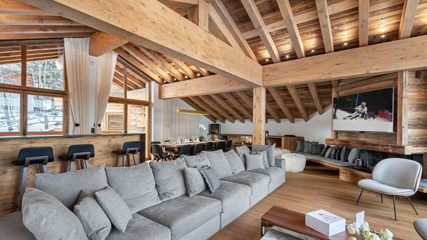 A spacious living area with a large grey sectional sofa, wooden beams, a television mounted on the wall, and a dining area in the background. - Exclusive Chalet in Courchevel Village (Courchevel 1550)
