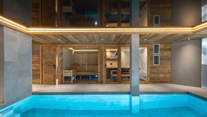 A modern indoor pool area with a clear view of the pool, steps leading into the pool, and a sauna area visible through glass doors. - Exclusive Chalet in Courchevel Village (Courchevel 1550)