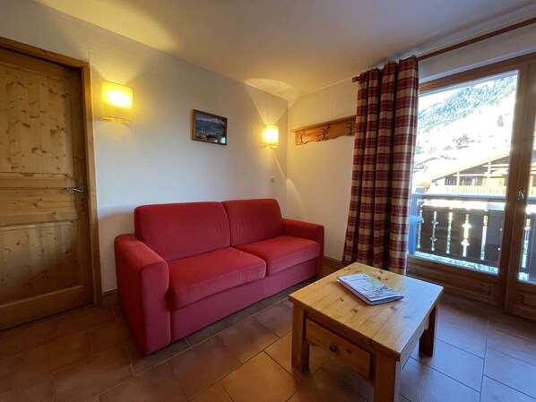 Ein rotes Sofa, ein Holztisch mit einer Zeitschrift, eine Holztür, ein Wandbild und ein Fenster mit Vorhängen.