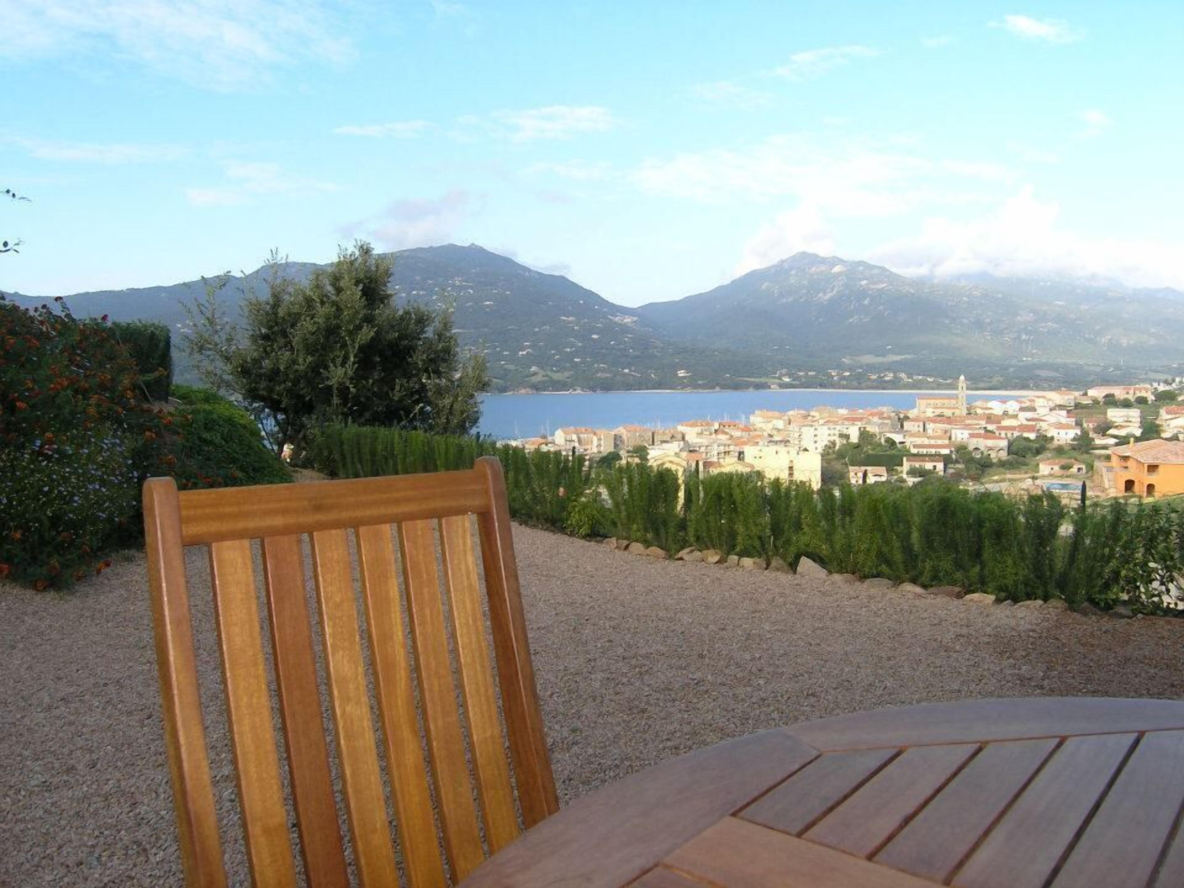 Una silla y una mesa de madera en un jardín con vistas a un pueblo costero y montañas.