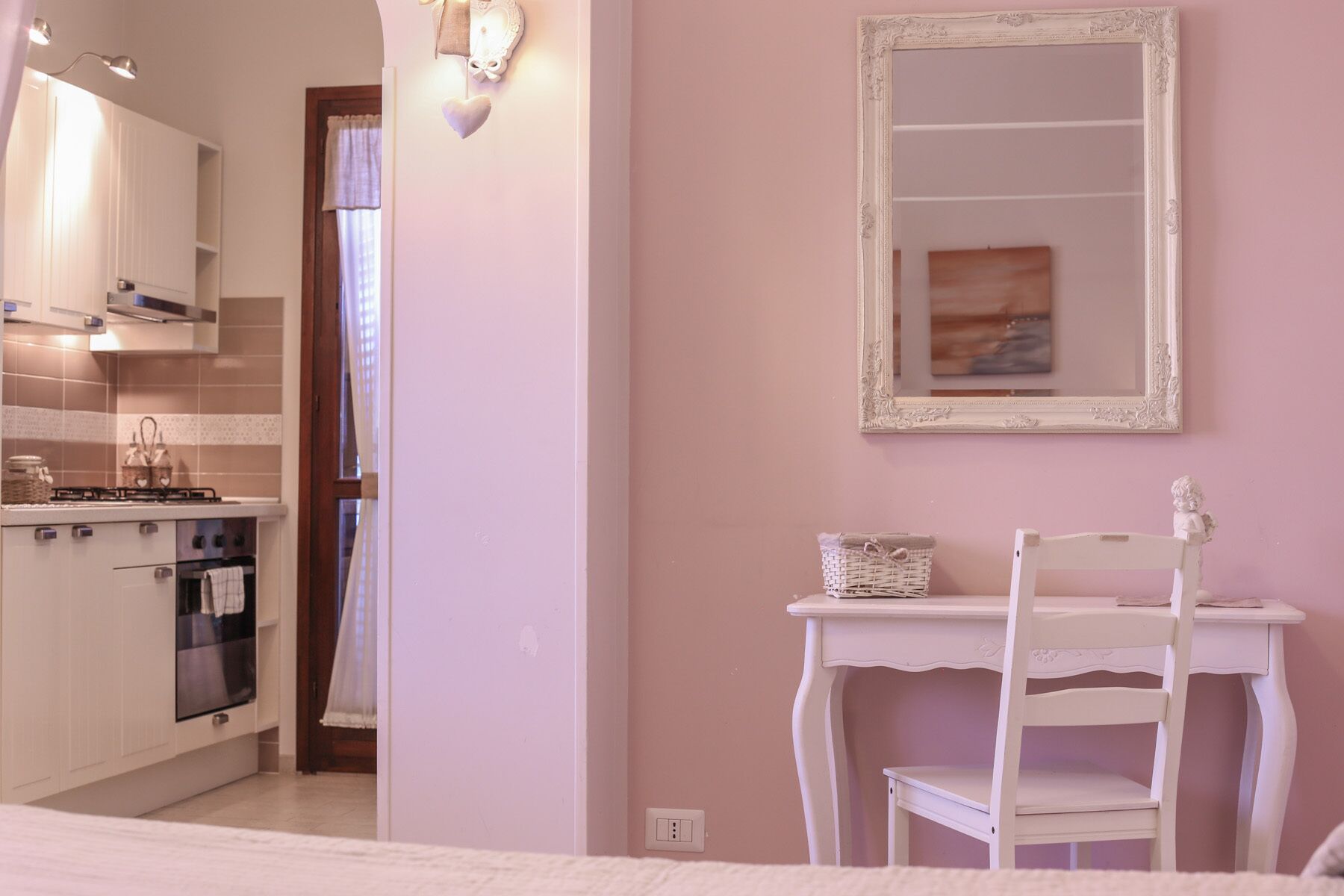 A small, cozy room with a kitchenette, a white chair, a small table, and a mirror on the wall.