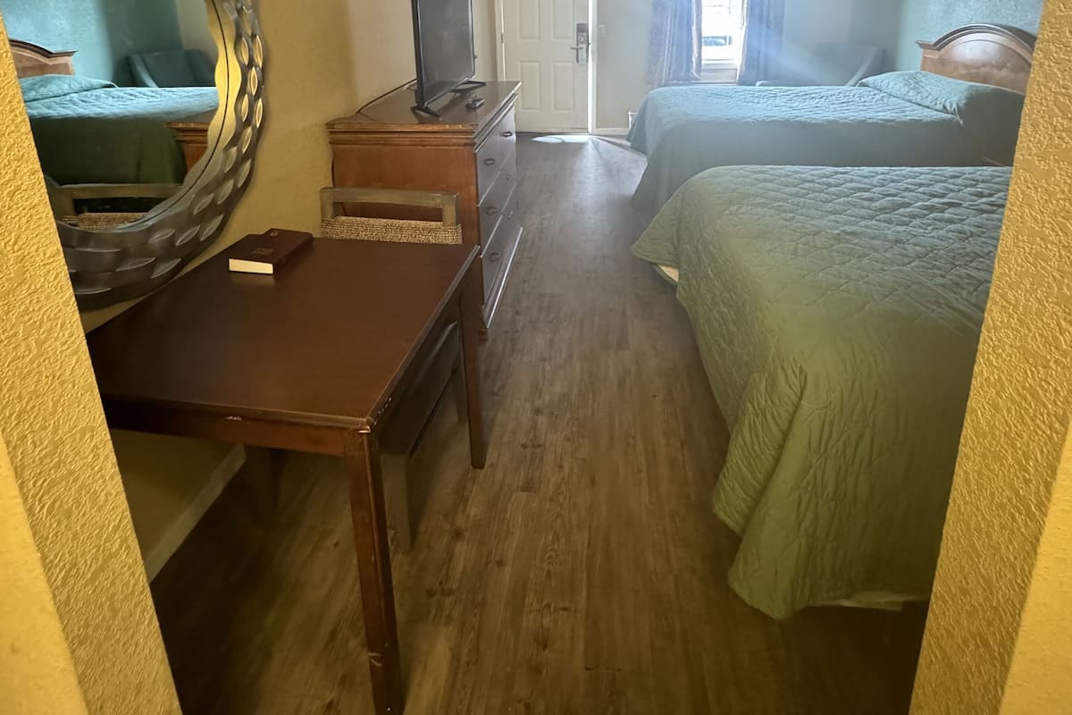 Habitación de hotel con un escritorio de madera, televisor, cama con ropa de cama verde y un espejo grande en la pared.