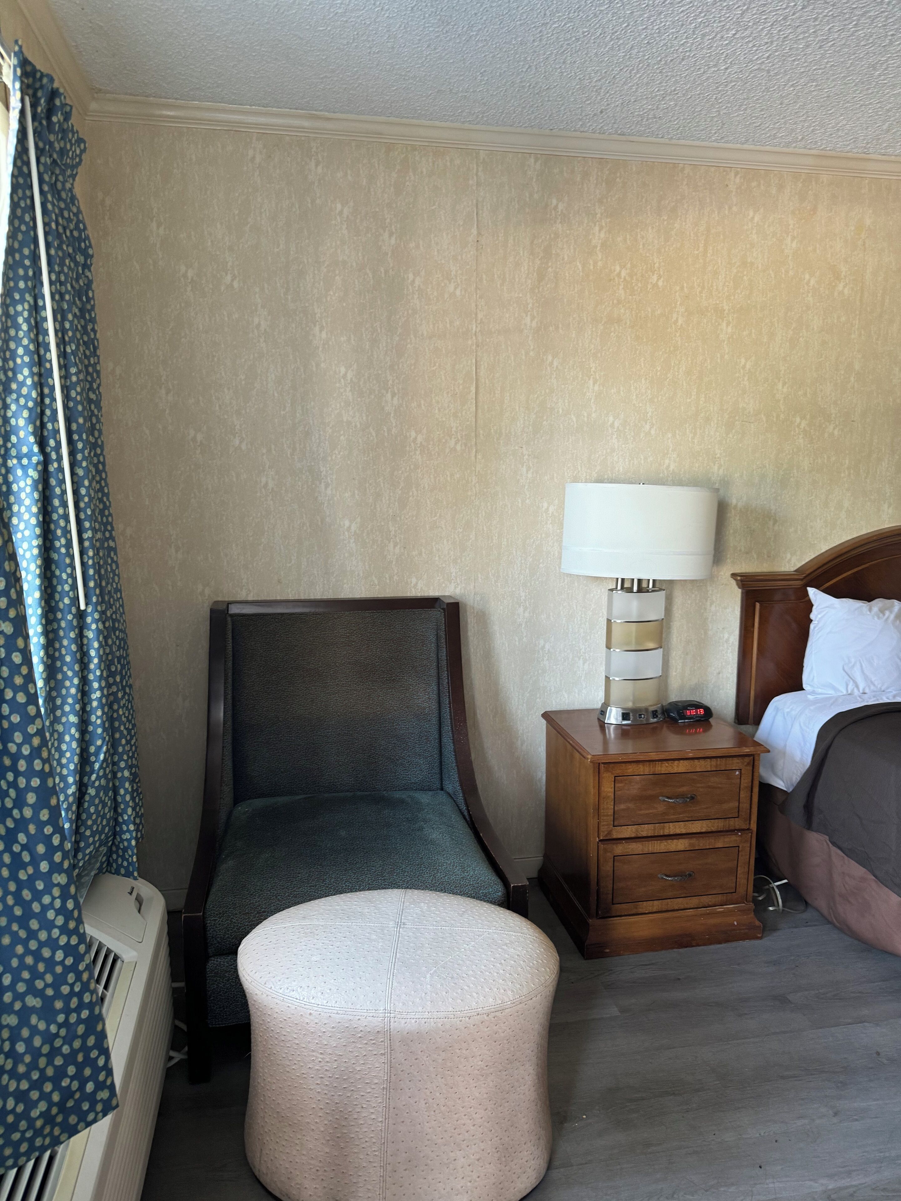 A hotel room with a single bed, a wooden nightstand with a lamp, a small round ottoman, a chair, and a curtain with a polka dot pattern.