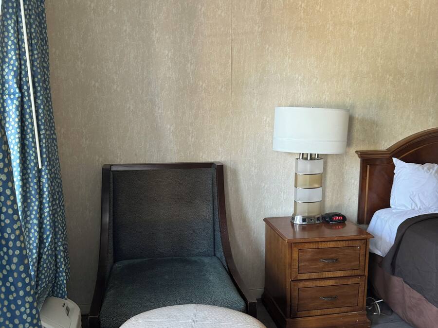 A hotel room with a single bed, a wooden nightstand with a lamp, a small round ottoman, a chair, and a curtain with a polka dot pattern.