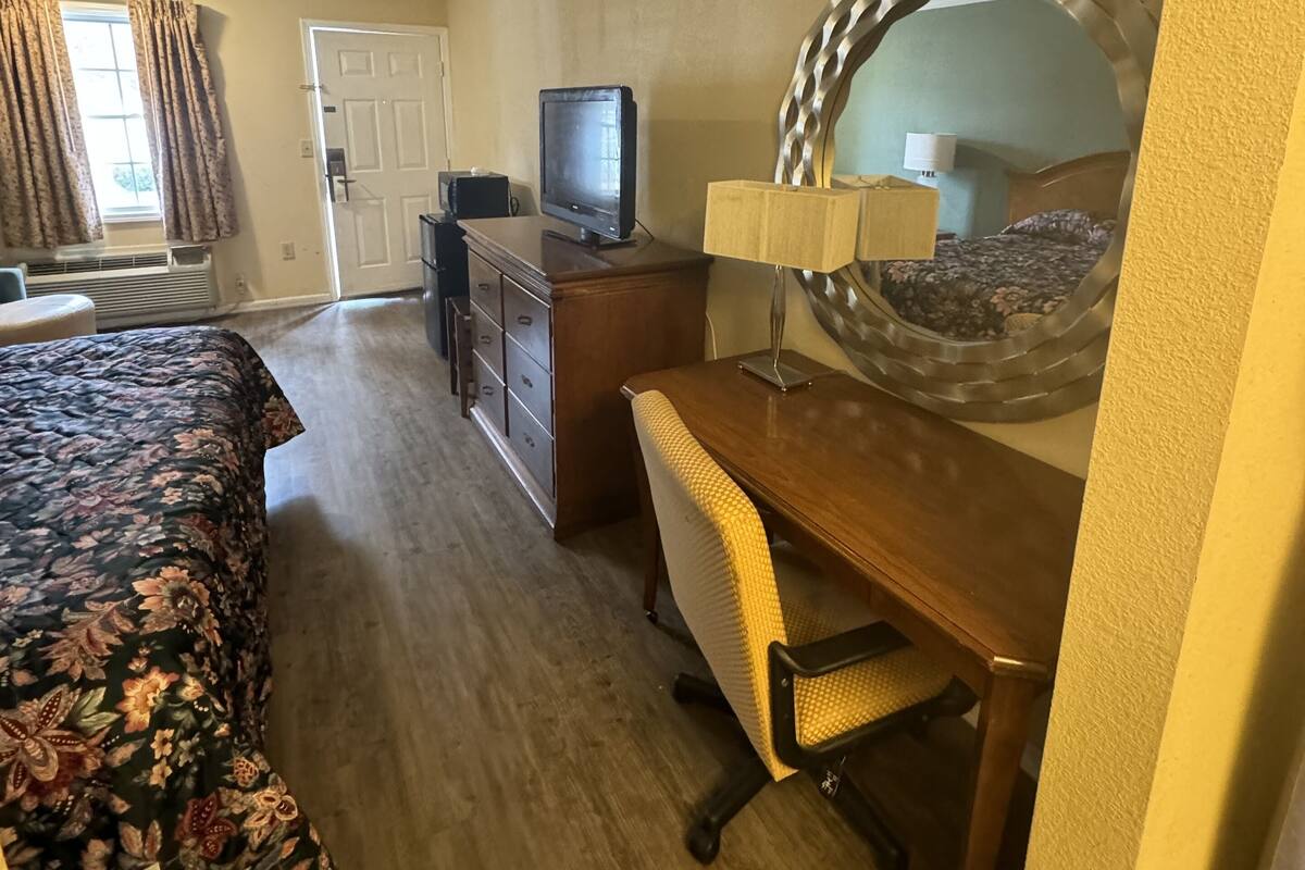 Habitación de hotel con cama, escritorio y silla, televisión, cómoda y un espejo grande.