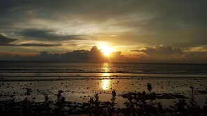Uma praia com o sol se pondo no horizonte, criando o contorno de nuvens e um mar tranquilo.