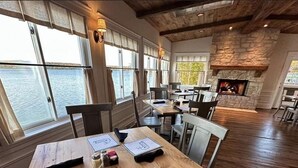 Una mesa de comedor de madera con tarjetas de lugar, una chimenea y grandes ventanales con vistas a un cuerpo de agua.