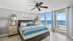 Un dormitorio con cama, mesitas de noche, ventilador de techo y vista al océano a través de amplias ventanas.