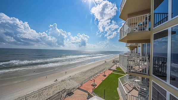 A beachfront hotel with a view of the ocean, a patio area with outdoor seating, and a grassy lawn.