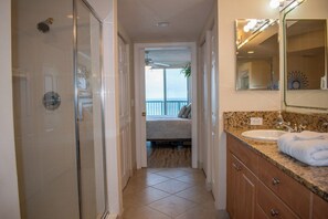Une salle de bain d’hôtel avec une douche en verre, un lavabo doté d’un plan de travail en granit et un lit visible à travers une porte ouverte.