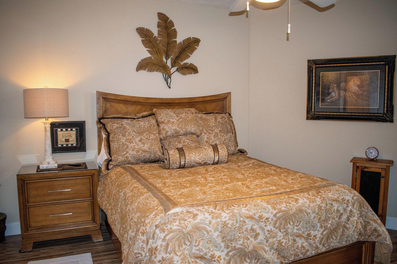Un dormitorio con una cama de madera, una mesita de noche con una lámpara, un cuadro en la pared y un reloj sobre la mesita de noche.