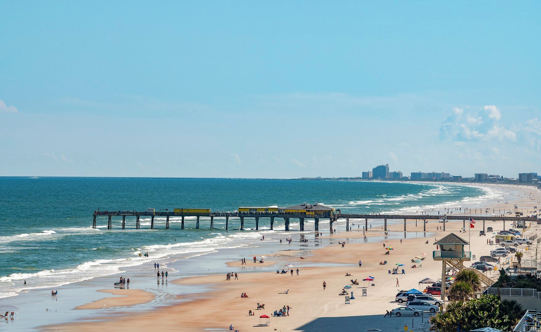 Una playa con un muelle que se adentra en el océano, arena y un horizonte urbano al fondo.