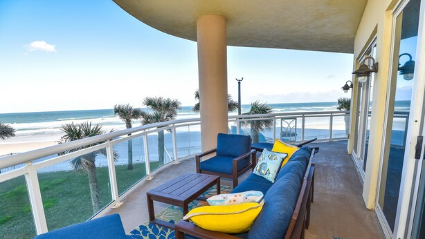 A balcony with a view of the beach, furnished with blue chairs and a table, and a patterned rug.