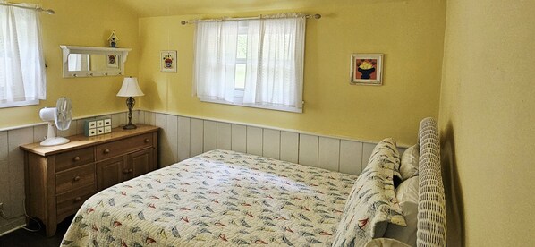A bedroom with a bed, a dresser, a mirror, a fan, and two windows with curtains.