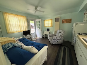 A compact room with a bed, a desk, a chair, a small kitchenette, and a ceiling fan.