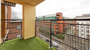 Un balcon avec un revêtement de gazon synthétique, une rambarde en métal et une vue sur un immeuble en briques rouges aux fenêtres vertes.