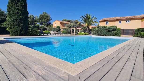 Una piscina rectangular con un muelle de madera que la rodea, ubicada en un área ajardinada con árboles y arbustos, y un edificio con techo de tejas al fondo.