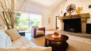 A living room with a sofa, armchairs, a coffee table with fruit, a fireplace, and a large window with a view of greenery. - Villa Lucendiluna. 10 pers, piscine, 5 min plage en voiture (Zonza)