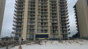 Un edificio residencial de varios pisos con balcones, el logotipo IV destacado y una playa arenosa en primer plano.