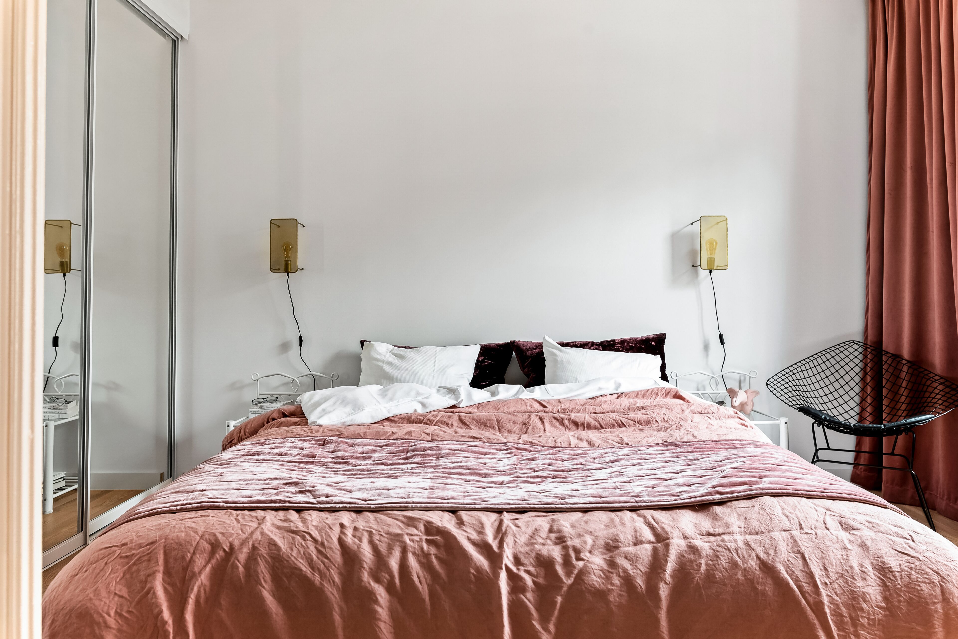 A neatly made bed with a pink bedspread, two bedside lamps, a black chair, and a glass wardrobe.