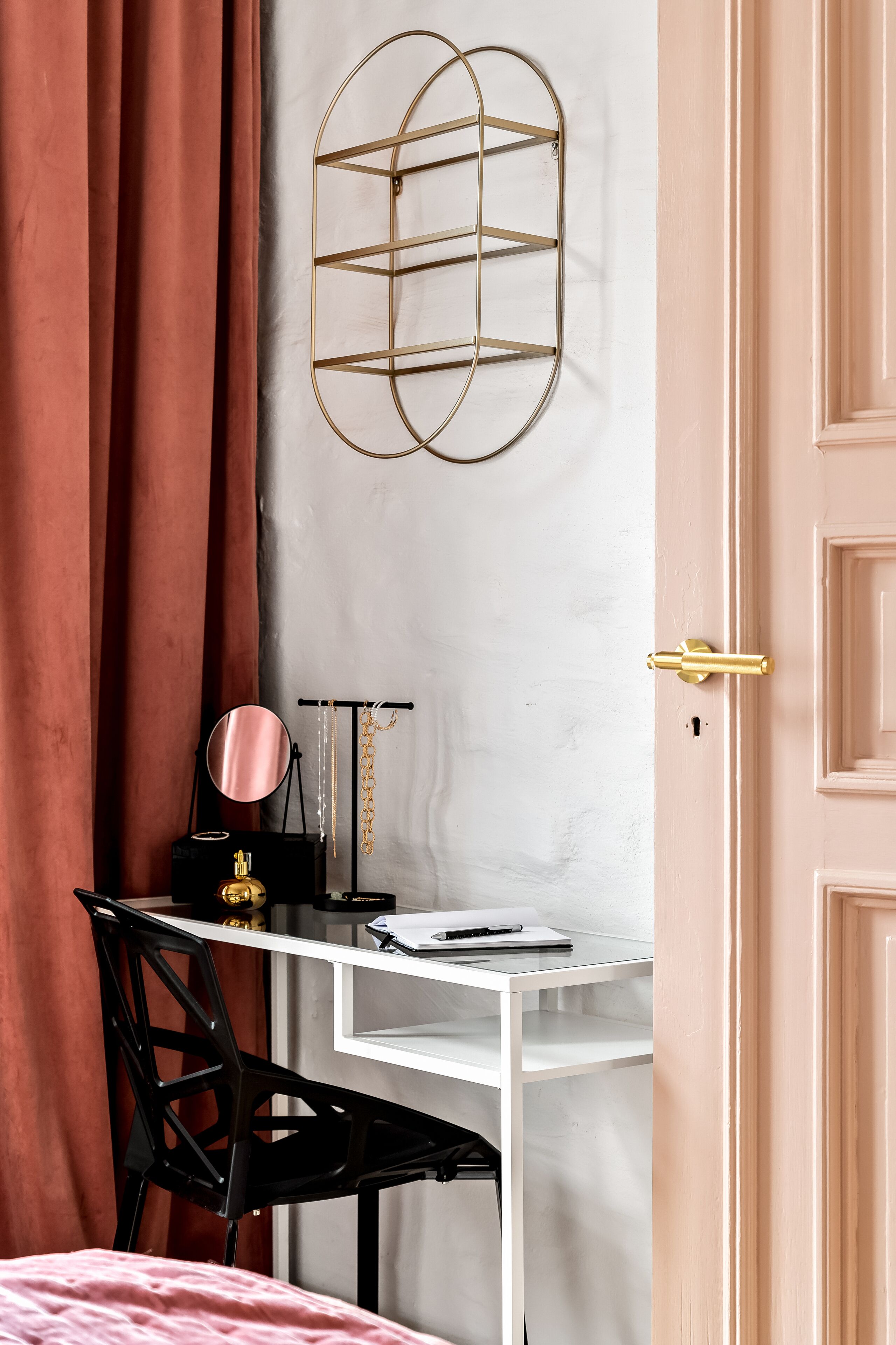 A room with a white desk, black chair, and a gold-framed mirror. A pink door is visible, and a black object with gold chains hangs on the wall.