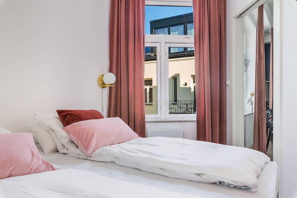 Un dormitorio con cama, almohadas rosas y blancas, una lámpara de pared y vistas a un balcón y un edificio a través de una ventana con cortinas rosadas.